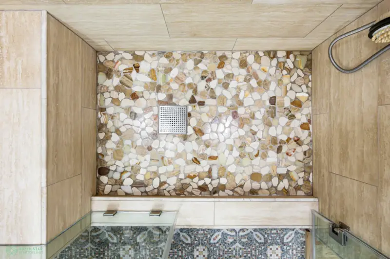 A top-down view of a walk-in shower featuring a natural pebble stone floor, a square stainless steel drain, and large beige travertine wall tiles.