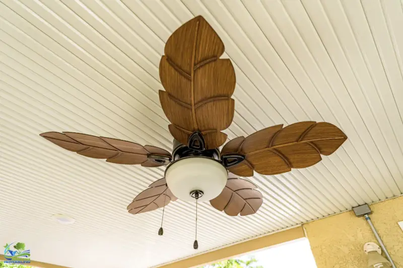 A ceiling fan featuring five wood-textured blades shaped like tropical leaves and a central frosted glass light fixture, installed on a white slatted patio ceiling.