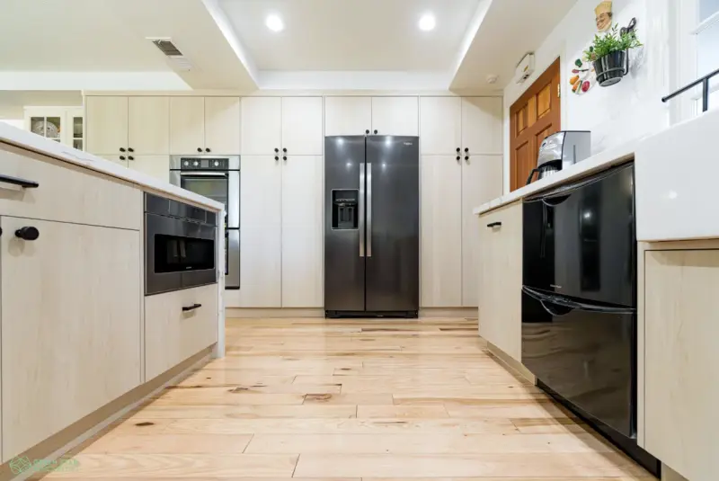 A modern kitchen featuring light-toned wood cabinetry, a large black stainless steel French door refrigerator, a built-in wall oven, and light hardwood floors.