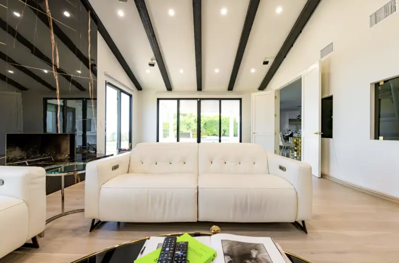 A bright, modern living room featuring a vaulted white ceiling with dark exposed wood beams and recessed lighting.