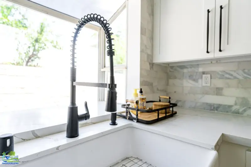 A modern matte black pull-down spring kitchen faucet installed on a white marble countertop with a large farmhouse sink in a remodeled kitchen.