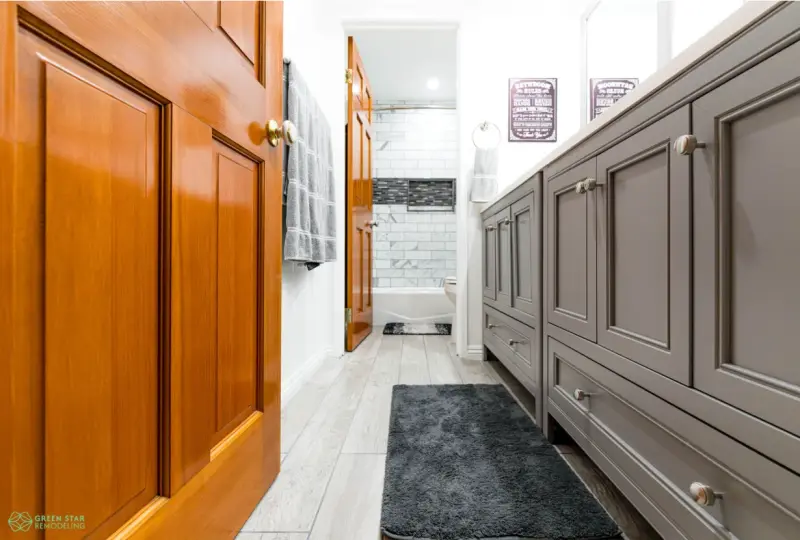 A modern bathroom interior featuring a dual gray shaker-style vanity with silver hardware, wood doors, light wood-look tile flooring, and a view into a remodeled shower area with marble-style tiling.