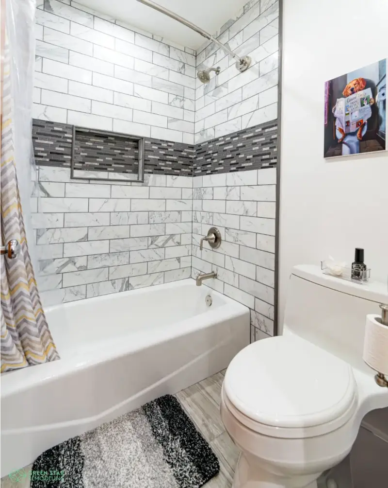 A remodeled bathroom featuring a white marble subway tile shower with a dark gray mosaic accent band, a white bathtub, and a modern white toilet.
