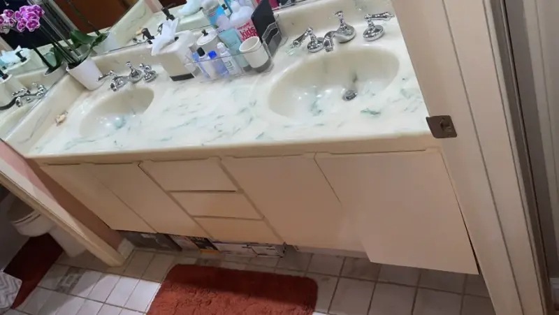 A double vanity bathroom featuring two sinks with chrome faucets, a marbled cream and green countertop, and cream-colored under-sink cabinets.