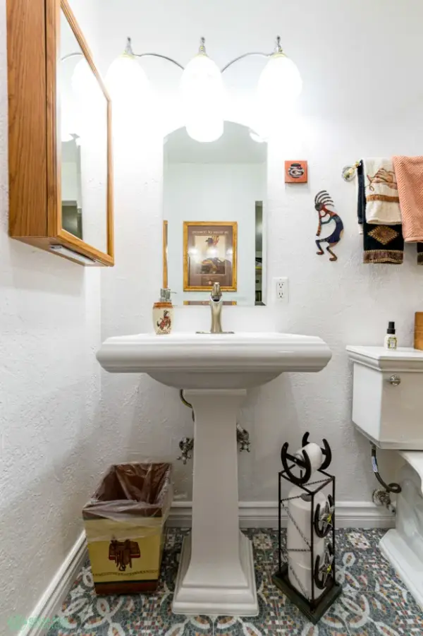 A brightly lit bathroom remodel in Calabasas featuring a white pedestal sink, patterned floor tiles, and Western-themed decor, including a horseshoe toilet paper holder and Kokopelli wall art.
