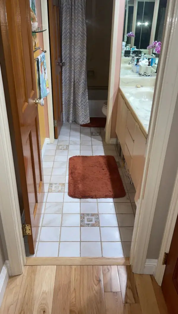 A view through a doorway into an old bathroom before a remodel, featuring light-colored square floor tiles with decorative accents, a wooden door, and a long cream-colored vanity with a marble-patterned countertop.