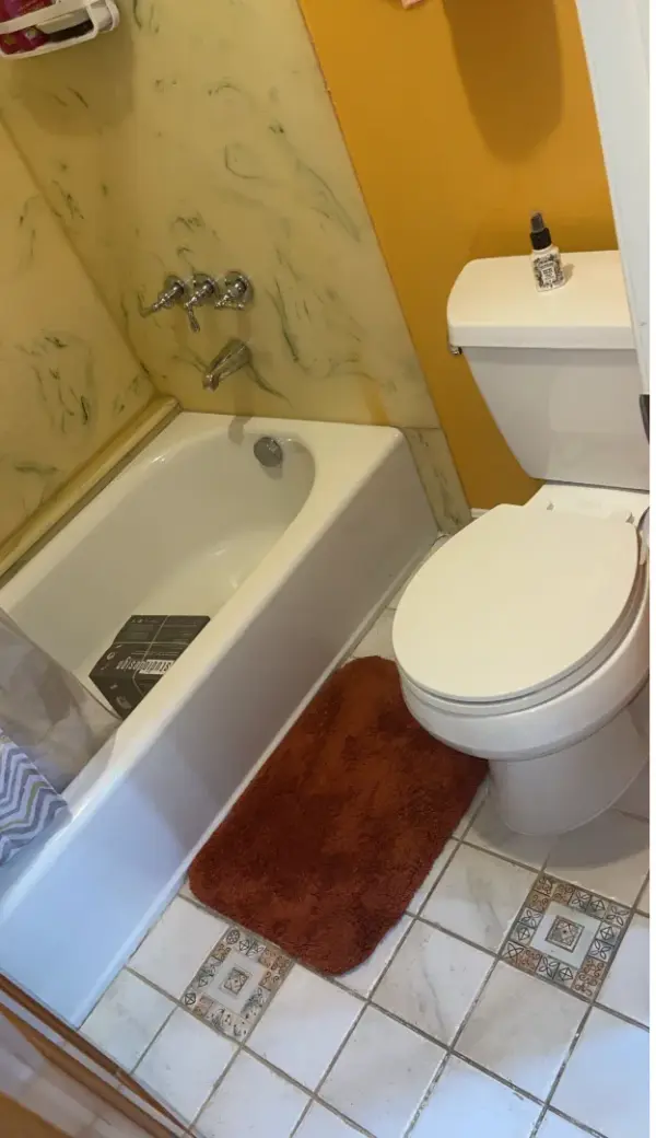 A small bathroom featuring a white bathtub and toilet, yellow walls, and a reddish-brown bath mat on a white tiled floor with decorative accents.