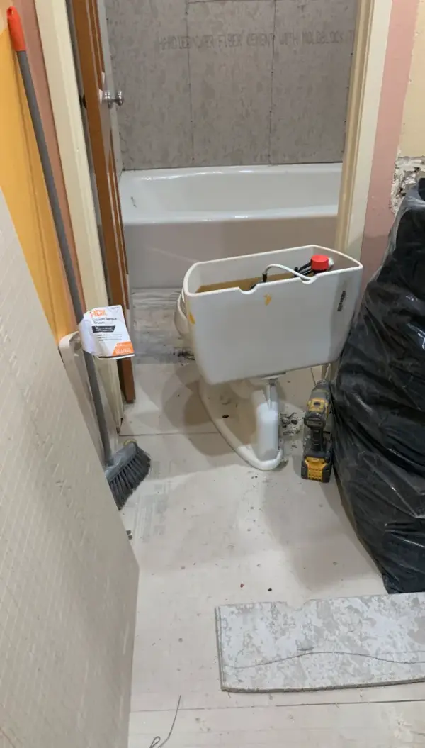 A bathroom undergoing renovation with a white bathtub and HardieBacker fiber cement board with Moldblock installed on the walls. A white toilet tank is sitting on the floor next to a yellow power drill and a black trash bag during the demolition or installation process.