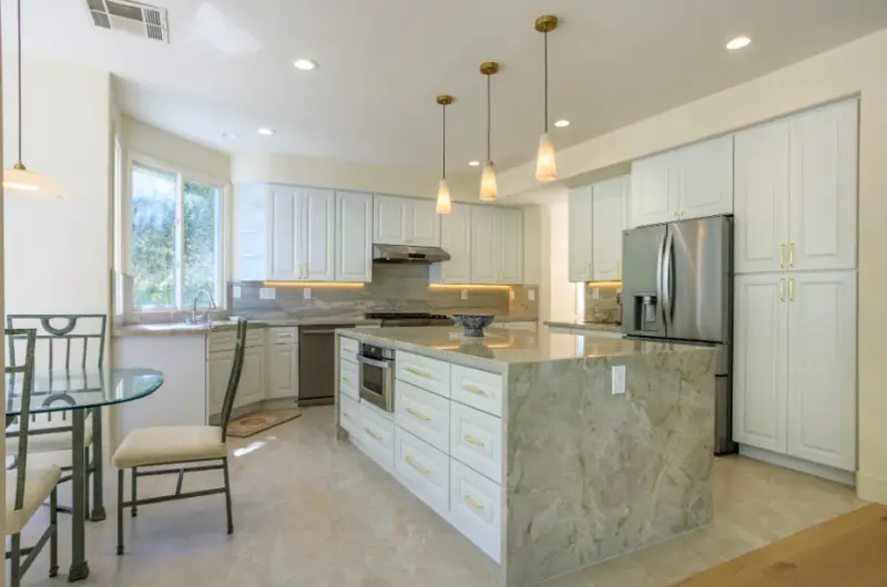 A modern kitchen featuring white cabinetry with gold hardware, a large marble waterfall island with an integrated oven, stainless steel appliances, and three pendant lights hanging over the center.