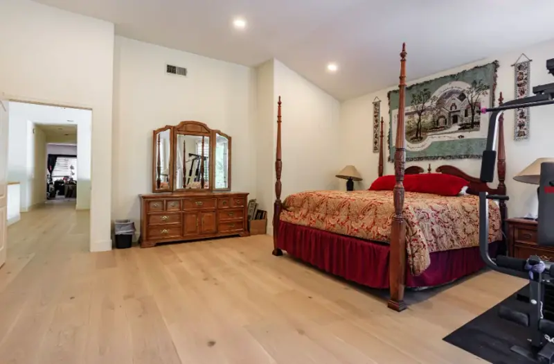 A spacious master bedroom featuring a tall wooden four-poster bed with red bedding, a matching wood dresser with a triple mirror, and light-toned hardwood flooring.