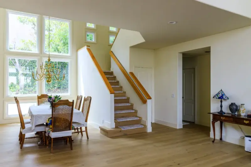 A bright and airy open-concept dining room featuring a wooden table with white chairs, a brass chandelier, and a wood-tread staircase with white banisters leading to the second floor, all set on light hardwood flooring.
