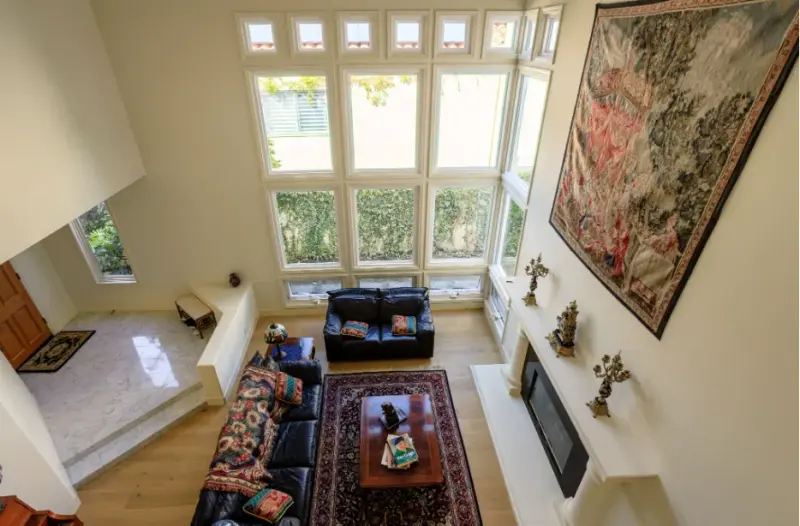 High-angle view of a bright living room featuring a double-height ceiling, a large grid of windows overlooking a garden, two dark leather sofas, a traditional rug, and a white fireplace mantle decorated with ornate candelabras.