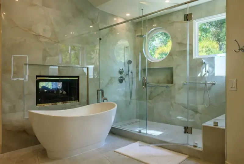 A modern luxury bathroom featuring a white freestanding soaking tub, a large glass-enclosed walk-in shower with marble tile walls, a circular accent window, and a built-in fireplace.