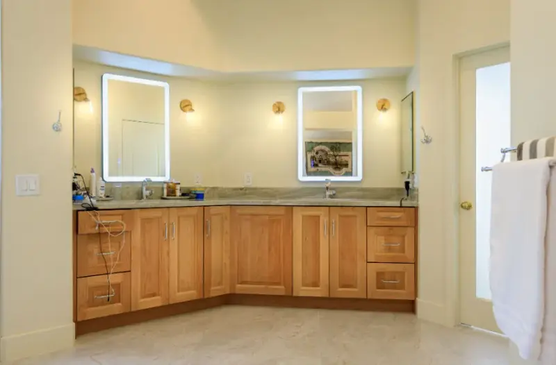 A remodeled bathroom featuring a curved double vanity with light wood shaker cabinets, a stone countertop, and two backlit rectangular mirrors framed by wall sconces.
