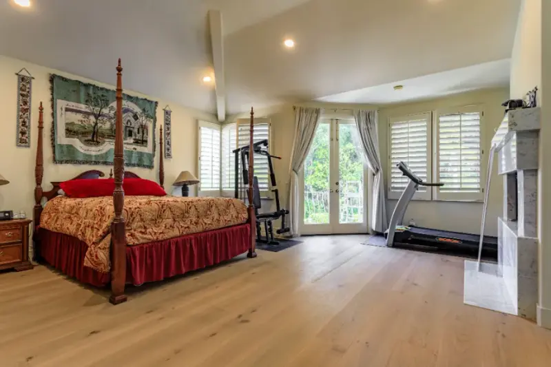 A spacious master bedroom featuring a four-poster wooden bed with red linens, a treadmill, a weight bench, and a white marble fireplace on light wood flooring.