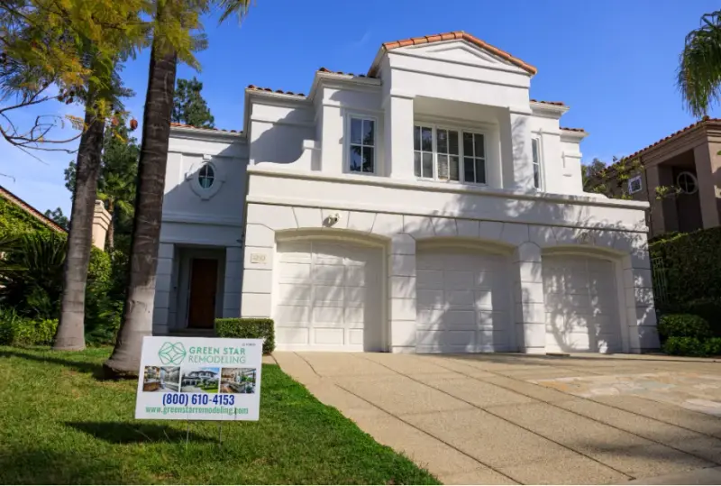 Front exterior view of a white, Mediterranean-style two-story luxury home in Calabasas featuring a three-car garage, red tile roof, a Green Star Remodeling yard sign, and surrounded by palm trees and lush landscaping.