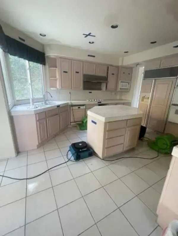 A kitchen undergoing water damage restoration with professional air movers and dehumidifiers placed on a white tile floor to dry light pink cabinets and a kitchen island.