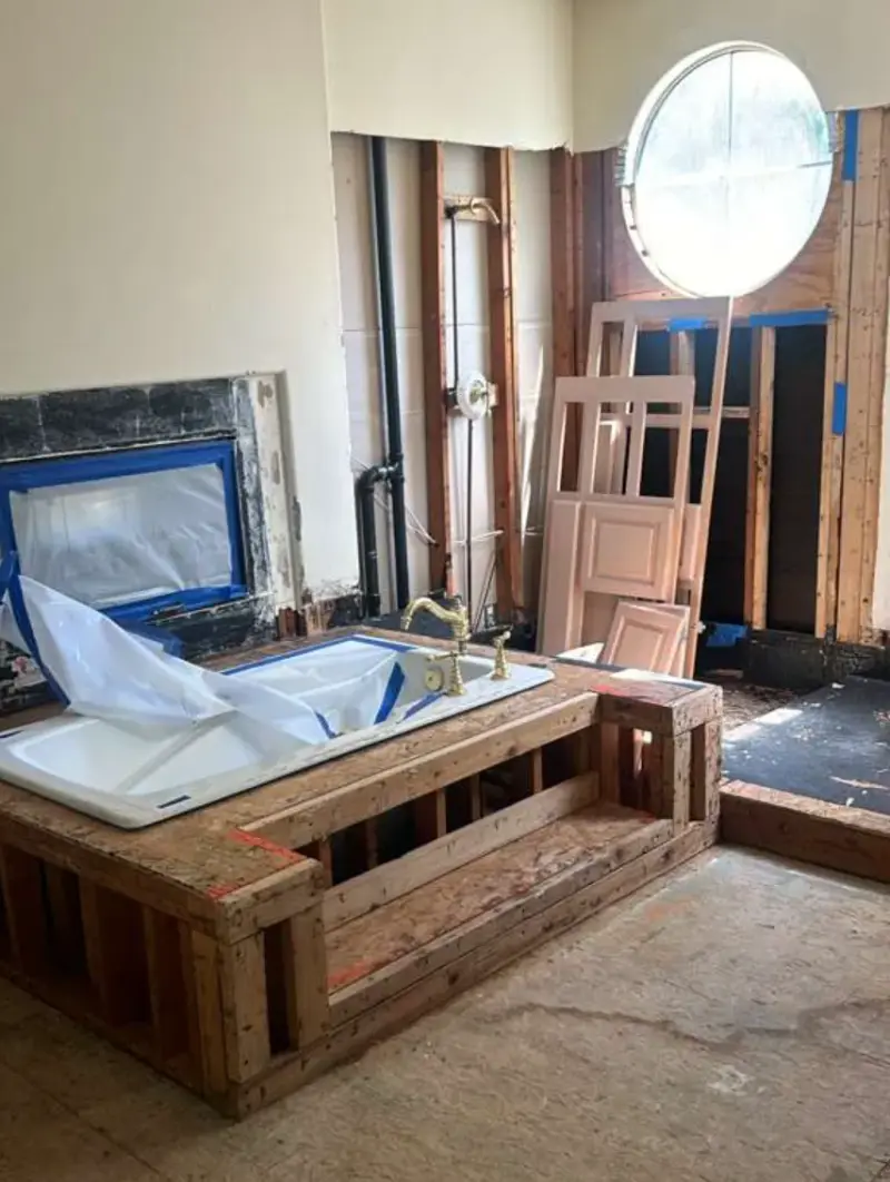 A bathroom undergoing renovation, featuring a white bathtub set into a newly constructed wooden frame with a step. The background shows exposed wall studs, plumbing pipes, and a circular window.
