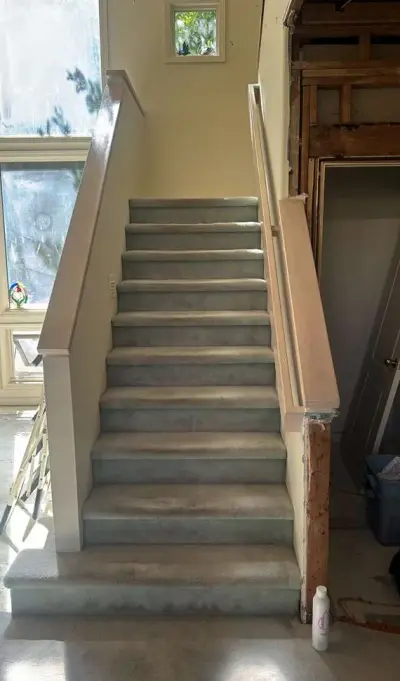 A staircase mid-renovation with bare concrete treads, unfinished handrails, and protective coverings on the surrounding walls and landing.