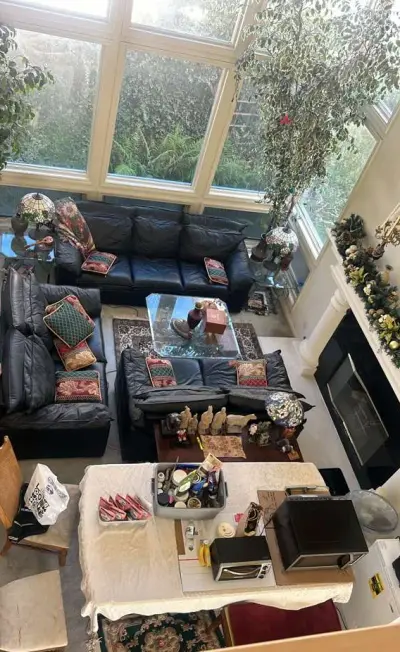A high-angle view of a sunlit living room featuring black leather sofas with patterned throw pillows, a large window wall overlooking greenery, a fireplace with a decorative mantel, and a dining table in the foreground.