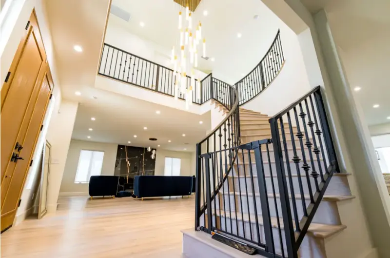 A bright, modern home entryway in Encino featuring a curved wooden staircase with black wrought iron railings, a safety gate, and a large multi-pendant chandelier hanging from a high ceiling.