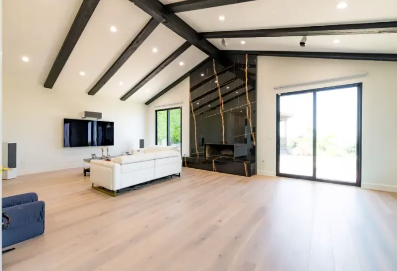 A modern living room featuring a vaulted ceiling with dark exposed wood beams, light hardwood floors, and a floor-to-ceiling black marble fireplace with gold veining.