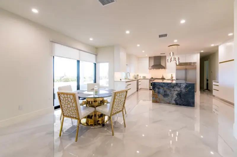 A modern luxury kitchen and dining area featuring a large blue marble waterfall island, white cabinetry, gold-accented chairs, and polished marble floors.
