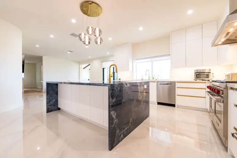 Modern luxury kitchen featuring a large blue marble waterfall island, white minimalist cabinetry with gold hardware, high-end stainless steel appliances, and polished marble flooring.