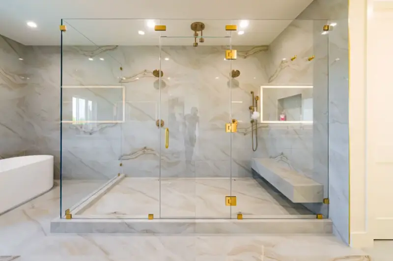A modern master bathroom featuring a large walk-in glass shower with gold hardware, marble-patterned wall tiles, a built-in bench, and dual shower heads next to a white freestanding bathtub.