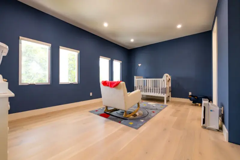 A modern nursery featuring navy blue walls, light-toned hardwood floors, a white wooden crib, and a cream-colored rocking chair on a celestial-themed area rug.