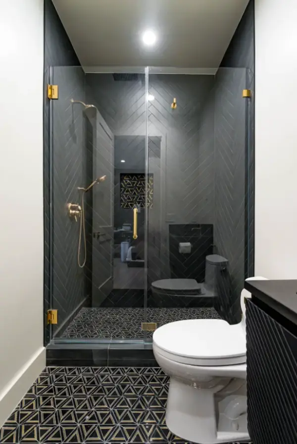 Modern bathroom remodel featuring a walk-in shower with dark grey herringbone wall tiles, gold hardware, and geometric patterned floor tiles next to a white toilet.