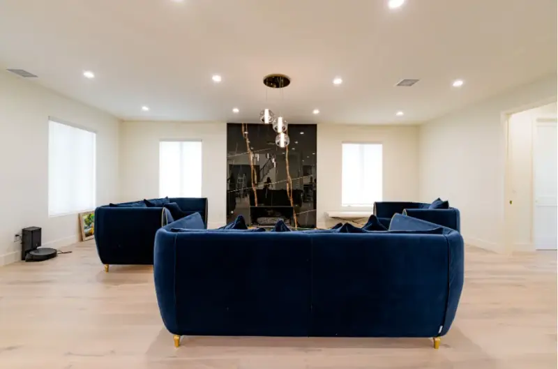 A spacious, modern living room featuring three deep navy blue velvet sofas with gold legs arranged around a sleek black marble accent wall with a built-in fireplace.