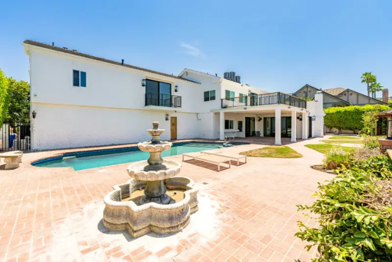 Rear view of a renovated two-story white home in Encino featuring a large swimming pool, a tiered stone water fountain, and a spacious brick patio with lounge chairs.