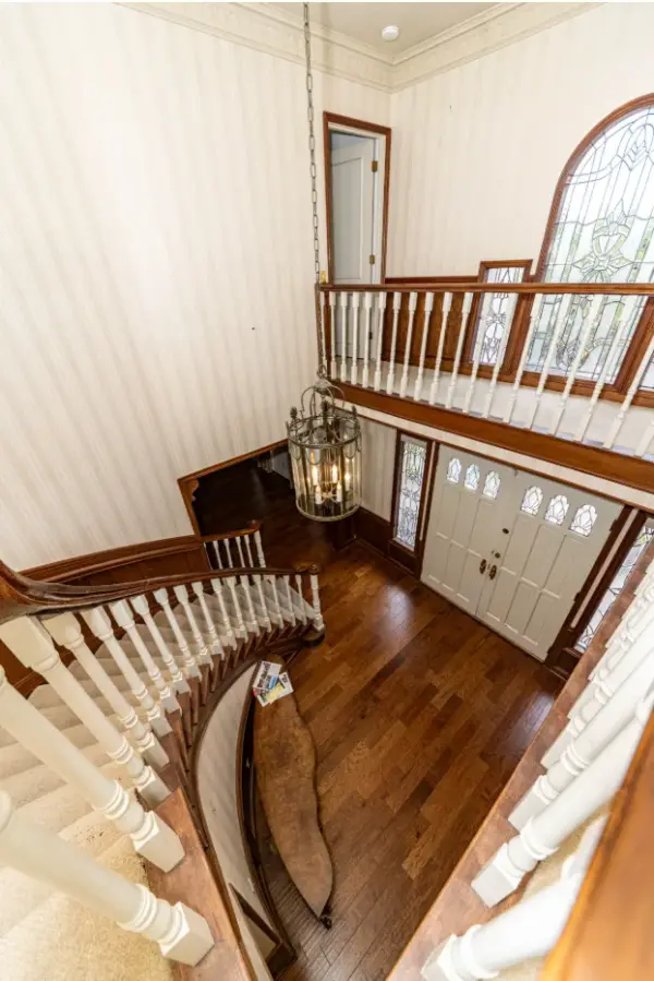High-angle view of a grand home foyer featuring a curved carpeted staircase with white balustrades, dark hardwood flooring, a hanging lantern chandelier, and white double entry doors with decorative glass panels.
