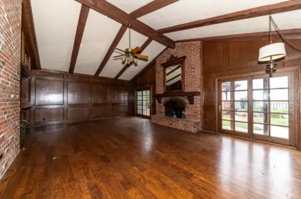 A large, empty living room featuring a vaulted ceiling with dark wood beams, dark wood-paneled walls, a brick fireplace with a mantle, and polished hardwood floors.