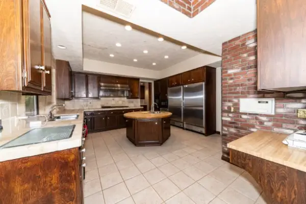 A spacious luxury kitchen featuring dark wood cabinetry, a circular kitchen island with a butcher block top, stainless steel double refrigerators, and a rustic red brick accent wall.