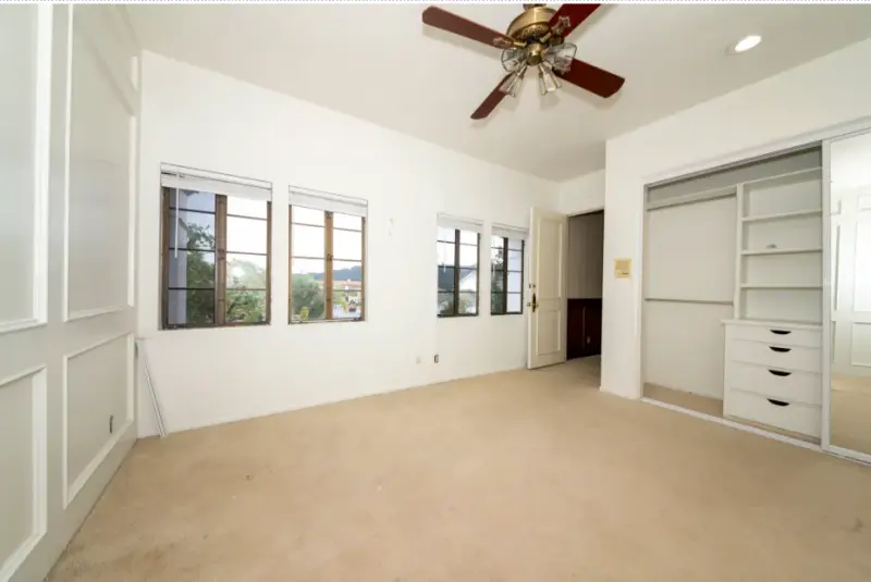 An empty bedroom with beige wall-to-wall carpet, white wainscoted walls, a dark wood ceiling fan, and a built-in closet with mirrored sliding doors and drawers.