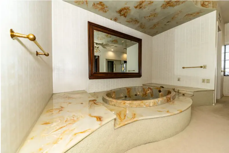 A dated bathroom featuring a curved onyx-look sunken tub surround, textured white wall tile, a wood-framed mirror with gold fixtures, and beige carpet flooring.