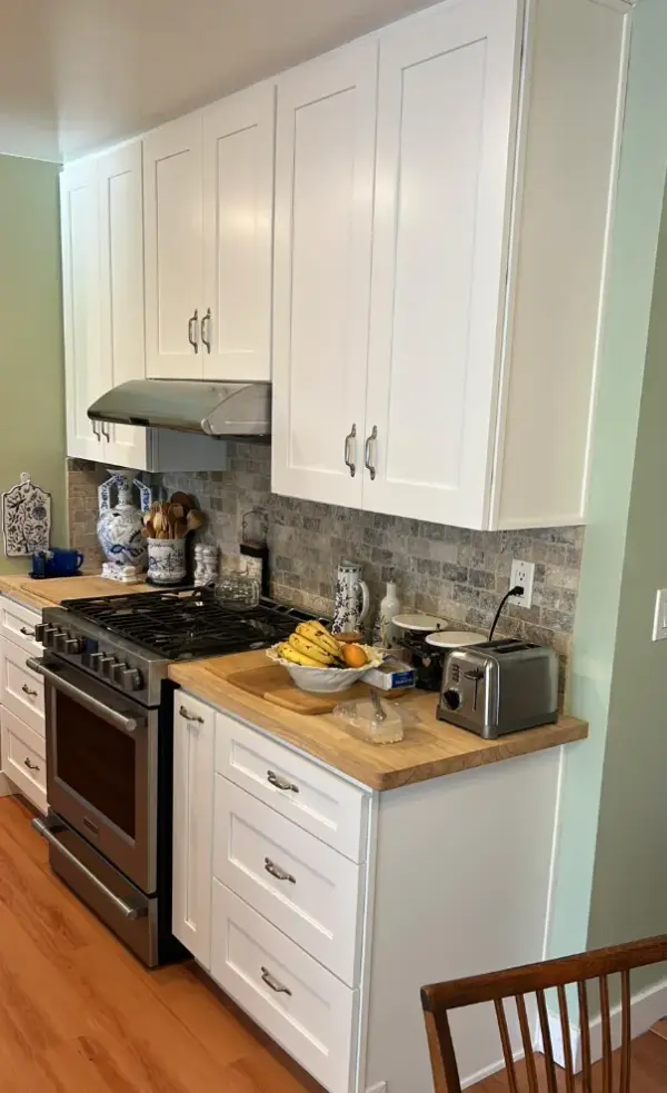 A remodeled kitchen featuring white shaker-style upper and lower cabinets, a light wood butcher block countertop, a stainless steel gas range with a matching vent hood, and a gray stone tile backsplash.