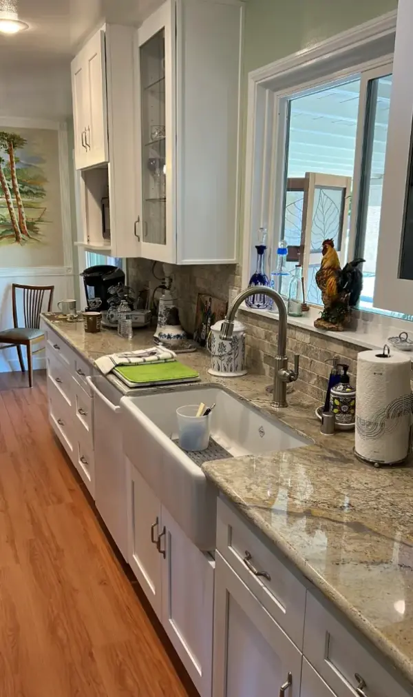 A remodeled kitchen featuring white shaker-style cabinets, light-colored granite countertops, and a white apron-front farmhouse sink.