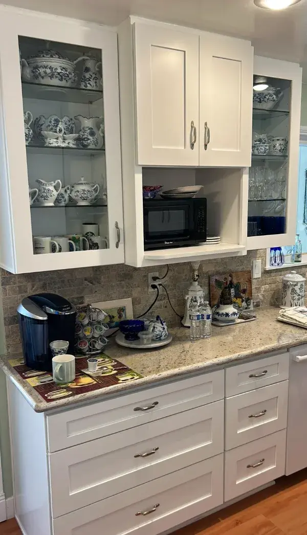 A remodeled kitchen featuring white shaker-style cabinets, glass-front upper cabinets displaying blue and white china, a black microwave built into a custom shelf, and speckled granite countertops with a stone tile backsplash.