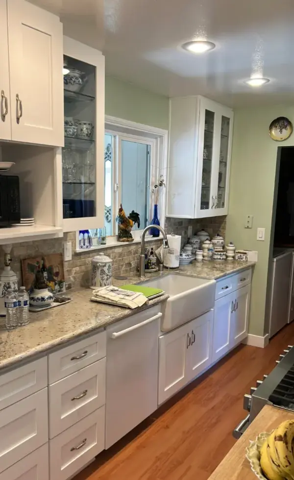 A remodeled kitchen featuring white shaker cabinets, a large white farmhouse apron sink, granite countertops, and light green walls.