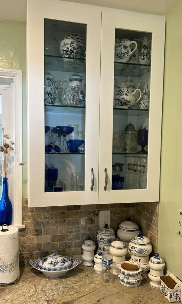 Modern kitchen remodel featuring white shaker-style glass-front cabinets, a neutral stone tile backsplash, and a collection of classic blue and white ceramic jars on a granite countertop.