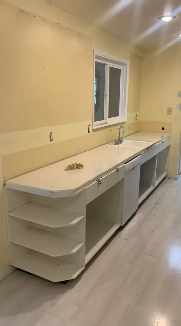 A long, white kitchen countertop with a built-in sink and dishwasher during a remodeling process, featuring open base shelving and pale yellow walls.