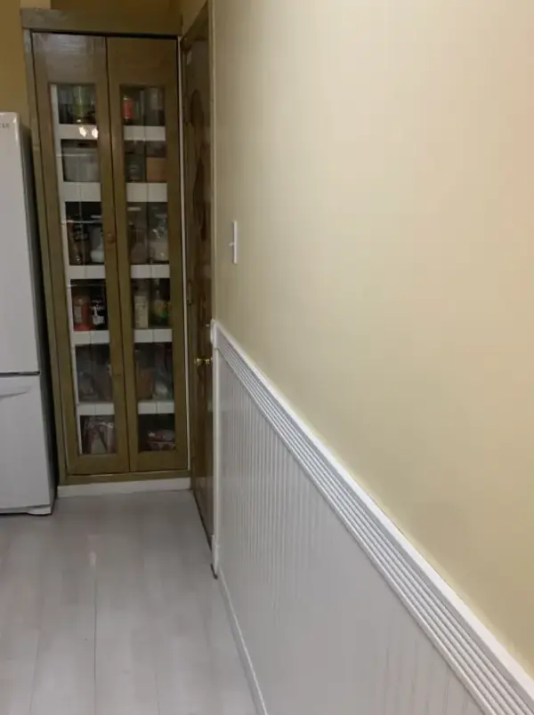 A kitchen interior featuring white beadboard wainscoting on a light yellow wall, light-colored wood-look flooring, a white LG refrigerator, and a tall wooden pantry cabinet with glass doors.