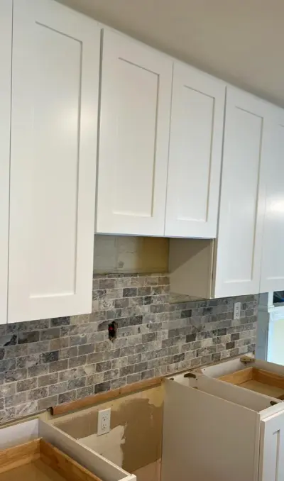 A kitchen undergoing a remodel featuring the installation of white shaker-style upper cabinets above a gray and tan stone tile backsplash.