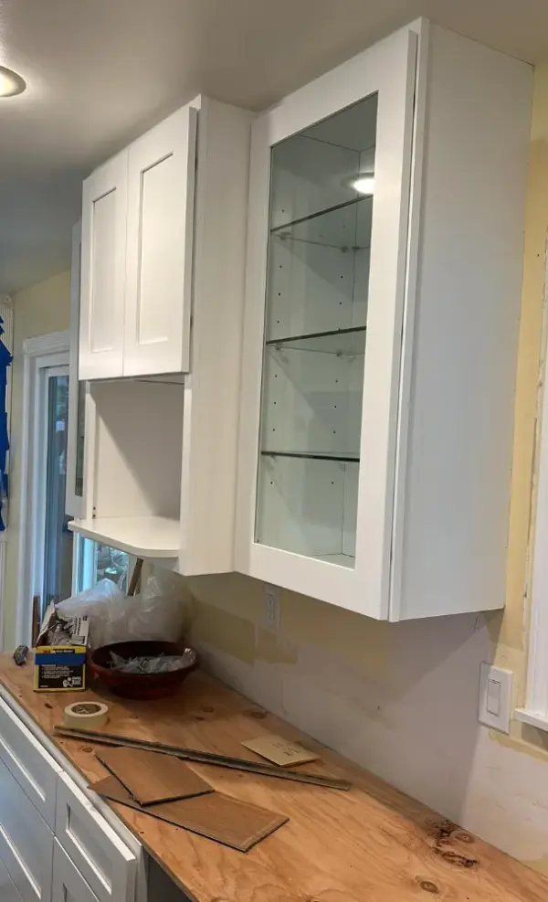 A kitchen remodeling project in progress showing newly installed white shaker-style upper cabinets, including one with a glass front and glass shelving, above a plywood countertop.