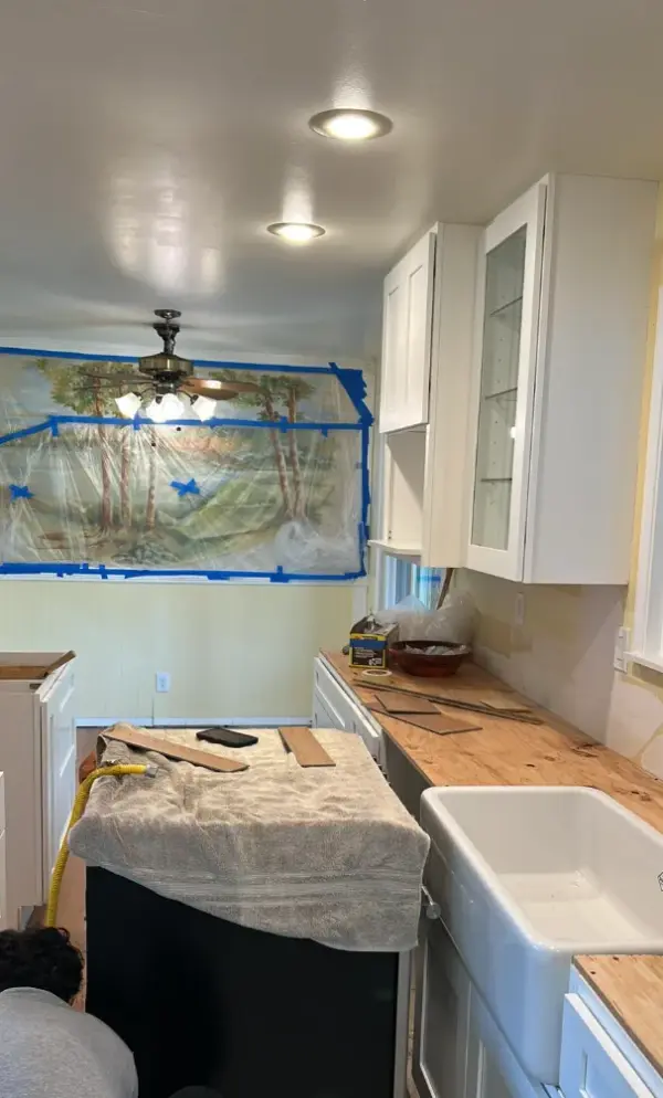 A kitchen undergoing a remodel featuring the installation of new white upper cabinets, a large white farmhouse apron sink, and a temporary plywood countertop next to an island.
