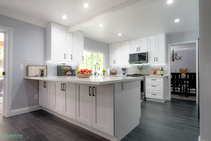 A modern kitchen remodel featuring white shaker cabinets, dark wood-style flooring, a marble-patterned countertop, and a white herringbone tile backsplash.