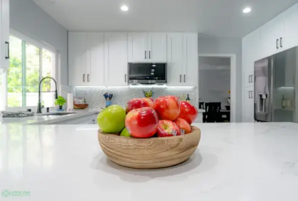 A modern kitchen remodel featuring white shaker cabinets, a white marble island with a wooden fruit bowl, and a herringbone tile backsplash by Green Star Remodeling.